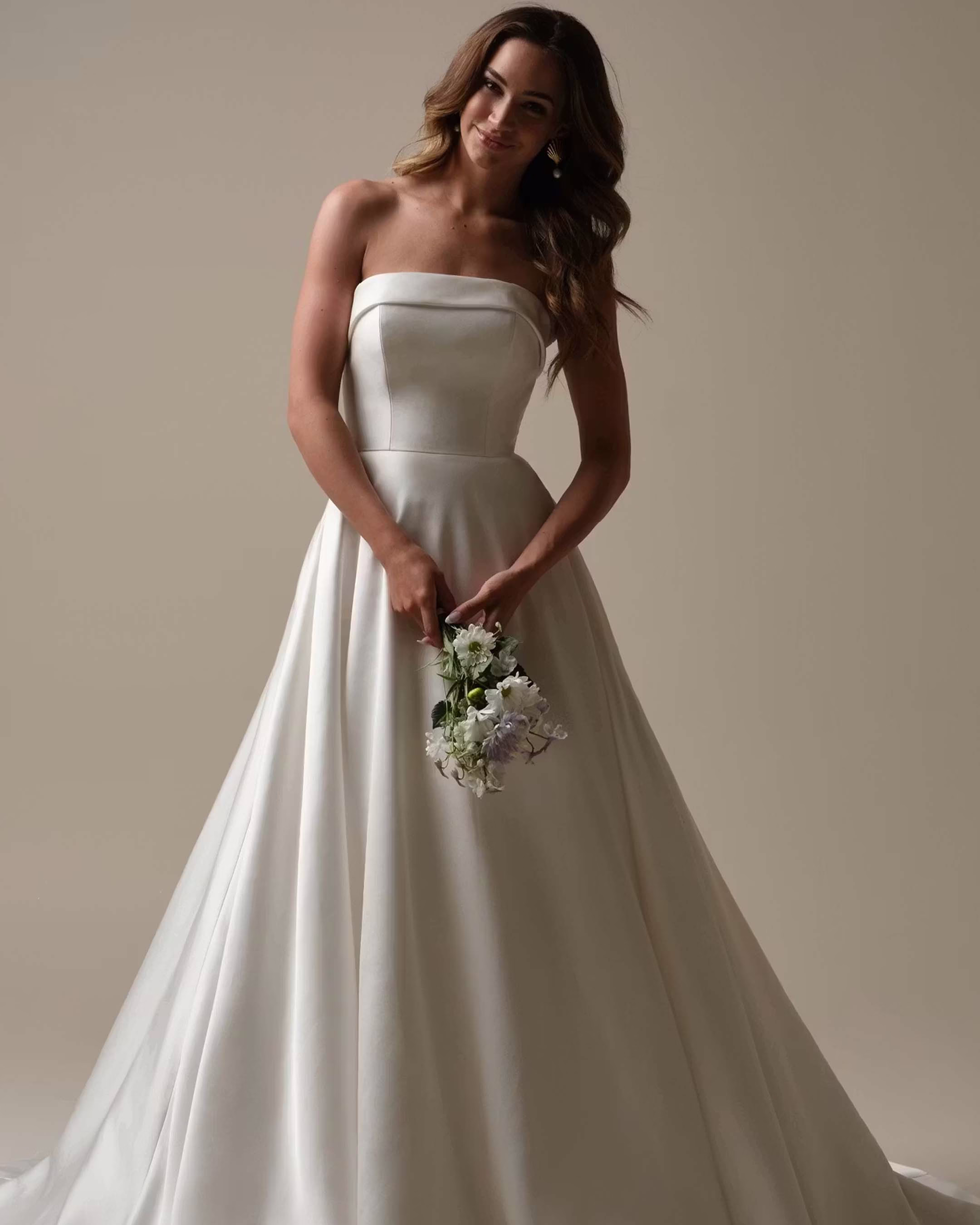 Rebecca Ingram Elizabeth Marie A-line wedding dress featuring a strapless satin bodice, banded cuff neckline, covered buttons, pockets, and elegant chapel train.