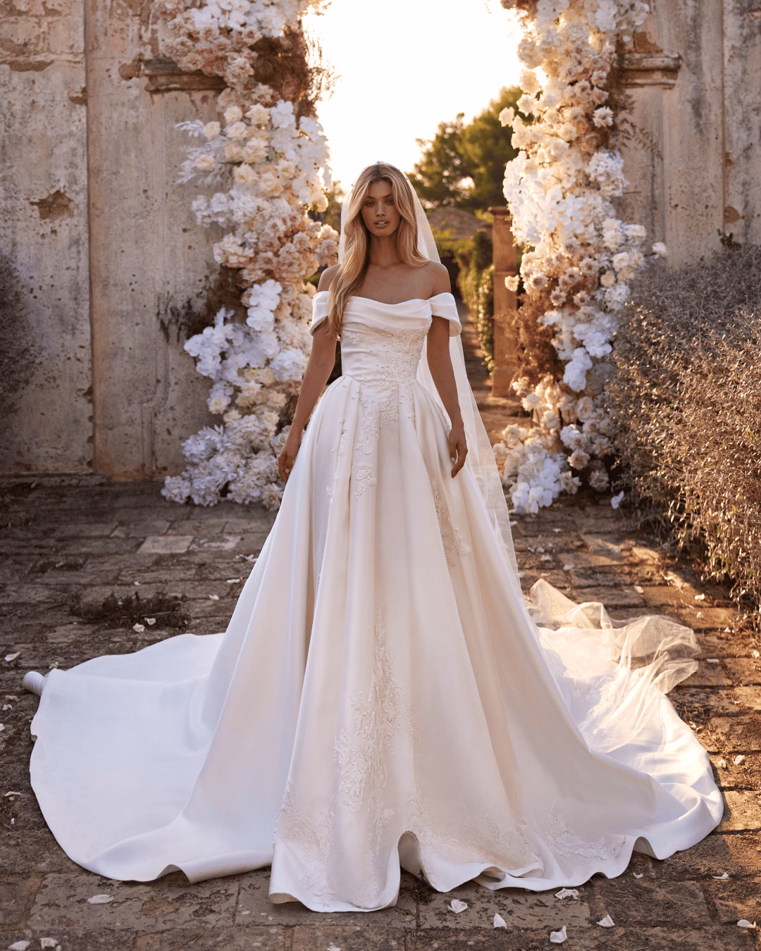 Milla Nova Symphony mikado princess wedding dress with an off-the-shoulder ruched corset bodice, hand-embroidered floral lace and sequins, full skirt, and cathedral train.